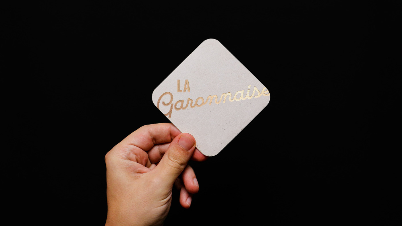 Sous-verre La Garonnaise au logo lettré en dorure, tenu à la main sur fond noir